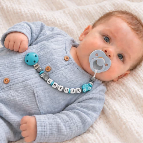 Bébé portant une attache-tétine personnalisée Arthur, avec deux petites voitures et des perles turquoise, argentées et gris foncé.